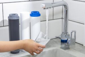 A person filling a glass of clear RO water from a faucet with three reverse osmosis filters sitting beside the sink